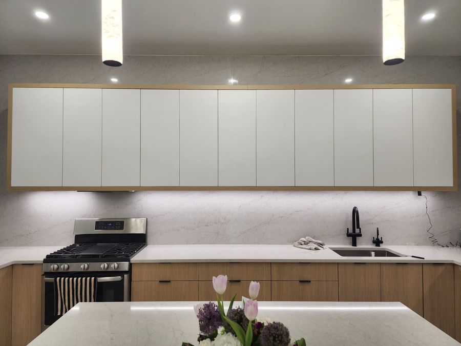 Wall of white flat-panel upper cabinets framed in oak over a white stone backsplash, oak lower cabinets, a black faucet and an island in front